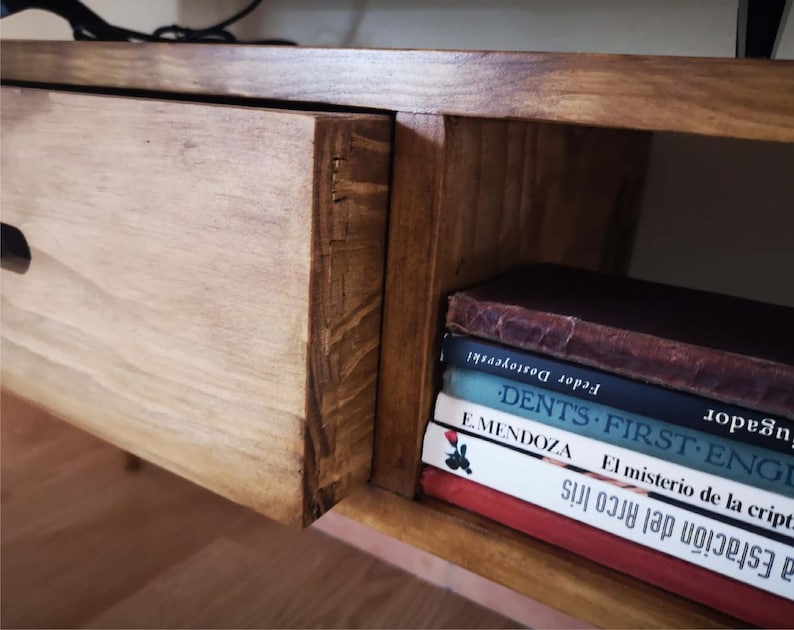May include: A wooden shelf unit with a drawer and a shelf. The shelf is filled with books, including "DENT'S FIRST ENGLISH" and "El misterio de la cript."