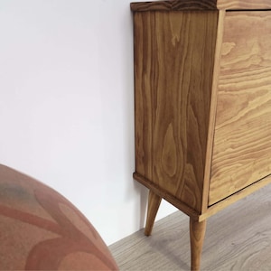 May include: A light brown wooden cabinet in a mid-century modern style. The cabinet has a drawer and sits on four tapered legs. The wood grain is visible. The cabinet is positioned against a white wall. The cabinet's dimensions are in centimetres.