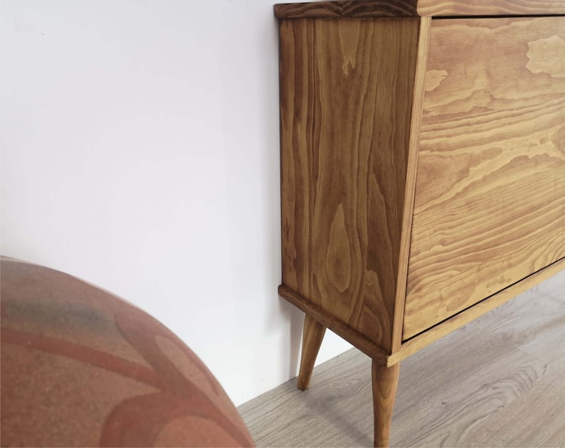 May include: A light brown wooden cabinet in a mid-century modern style. The cabinet has a drawer and sits on four tapered legs. The wood grain is visible. The cabinet is positioned against a white wall. The cabinet's dimensions are in centimetres.