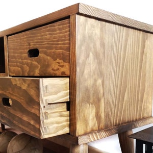 May include: A wooden entertainment center with a dark brown stain. The cabinet has three drawers and a shelf. The drawers have a simple, rectangular design with a small, rectangular cutout for a handle. The cabinet has a simple, rectangular design with rounded edges. The cabinet is sitting on four legs.