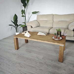Op de afbeelding: Een rechthoekige houten salontafel met een natuurlijke afwerking en vierkante poten. Op de tafel staan een bordje met cactussen, een open boek en een bril. Een beige bank en potplanten zijn op de achtergrond te zien.