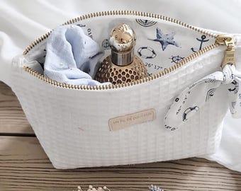 May include: White zippered pouch with a waffle-like texture. The pouch is open and contains a blue cloth, a gold bottle with a gold cap, and a white fabric with a nautical design. The fabric has a compass, anchor, and star.