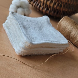 May include: A stack of white reusable makeup remover pads made of soft, textured fabric. The pads are folded and arranged on a wooden surface with a piece of twine.
