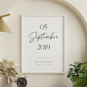 May include: A white framed print with black text that reads "05 September 2019" and "Michaela & Jack Marbella, Spain". The print is hanging on a white wall with a gold lamp and a vase of dried flowers on a shelf below.