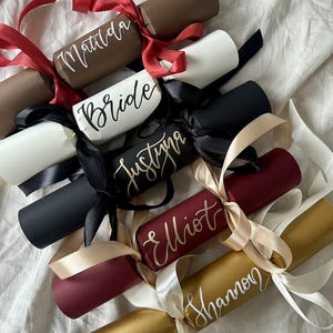 May include: A selection of colourful party crackers, each with a name written on it in elegant script. The crackers are in shades of brown, white, black, burgundy, and gold, and are tied with ribbons. Names include "Matilda," "Bride," and "Shannon."