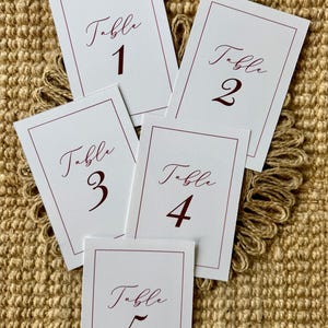 Peut inclure: Cinq cartes de table rectangulaires blanches avec une fine bordure bordeaux. Chaque carte présente le mot Table dans une police cursive au-dessus d'un grand chiffre, allant de 1 à 5 dans une police serif bordeaux, sur papier cartonné.