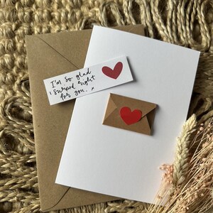 Könnte beinhalten: Eine weiße Karte mit einem roten Herzaufkleber und einem kleinen braunen Umschlag mit einem roten Herzaufkleber. Ein kleines weißes Rechteck mit dem Text "I'm so glad I swiped right for you." ist ebenfalls enthalten. Im Hintergrund ein brauner Umschlag.