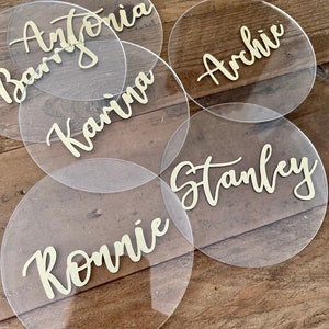Puede incluir: Cinco tarjetas de lugar redondas de acrílico transparente con letras doradas. Los nombres en las tarjetas de lugar son Antonia, Karina, Archie, Ronnie y Stanley.