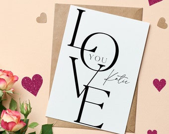 May include: A white card with the word "LOVE" written in black, with the word "YOU" inside the "O". The card is signed "Katie" and surrounded by pink glitter hearts on a light pink background.