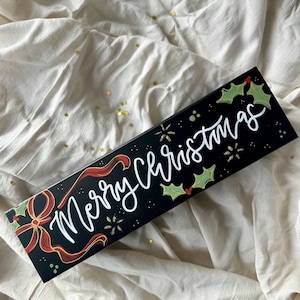 May include: A rectangular black Christmas-themed box with the words "Merry Christmas" in white script. The box features red ribbon and holly accents. Gold star-shaped confetti is scattered around the box.