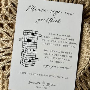 Puede incluir: Una tarjeta blanca con texto negro y una ilustración de una torre Jenga. El texto dice "Por favor, firme nuestro libro de visitas" e incluye instrucciones para los invitados. La tarjeta está firmada "Samantha & Stephen" con una fecha.