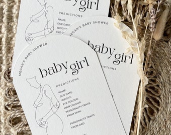 May include: Two white cards with a black line drawing of a pregnant woman on each. The cards are shaped like an arch and have the text "baby girl" and "predictions" on them. The text "Megan's Baby Shower" is on the top of the card.