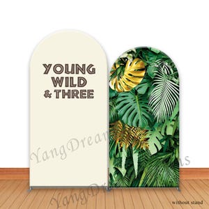 Peut inclure: Un fond de scène à deux panneaux avec un sommet en forme d'arche. Un panneau est de couleur crème avec le texte "YOUNG WILD & THREE" en marron. L'autre panneau présente un motif de feuilles tropicales vertes luxuriantes.