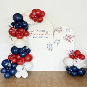 Puede incluir: Decoración festiva con globos rojos, blancos y azules. El arco de globos se complementa con un fondo blanco con el texto "A LITTLE FIRECRACKER IS ON THE WAY" y motivos de fuegos artificiales.