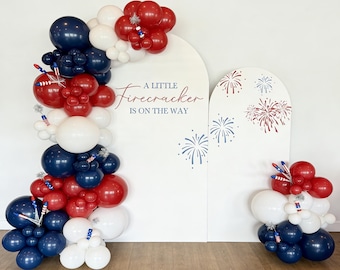 Puede incluir: Decoración festiva con globos rojos, blancos y azules. El arco de globos se complementa con un fondo blanco con el texto "A LITTLE FIRECRACKER IS ON THE WAY" y motivos de fuegos artificiales.
