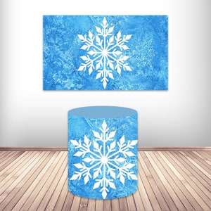 May include: A blue cylinder with a white snowflake design. The cylinder is sitting on a wooden floor. The background is a white wall with a blue rectangle with a white snowflake design.