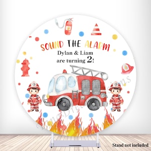 May include: A round backdrop with a watercolor design featuring a red and white fire truck, two cartoon firefighters, and the text "SOUND THE ALARM Dylan & Liam are turning 2!". The backdrop includes fire flames and fire-related illustrations.