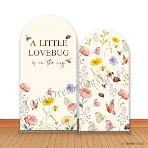 May include: Two arched backdrop banners with floral and insect designs. One banner reads "A LITTLE LOVEBUG is on the way." The other banner features flowers, butterflies, bees, and ladybugs. The banners are cream-colored with a wooden floor.
