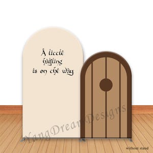 May include: A beige arch-shaped backdrop with the text "A little halfling is on the way" in black script. Next to it is a brown door with vertical lines and a circular detail. The backdrop and door are on a metal stand.