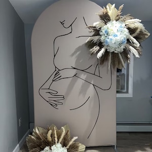 May include: A light brown backdrop with a black line drawing of a pregnant woman. The woman is standing with her hands on her stomach. There are dried pampas grasses and white and blue flowers on either side of the backdrop.