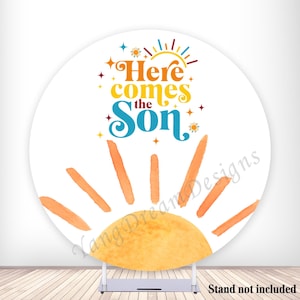 May include: A round white backdrop with a watercolor sun design. The sun has orange rays and a yellow center. Text in the center reads "Here comes the Son" in orange, yellow, and teal. Decorative stars and sun rays surround the text.