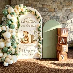 May include: An arched sign for a baby shower featuring a teddy bear reading a book, the text A NEW CHAPTER WILL SOON BEGIN and WELCOME TO Walker's BABY SHOWER, decorated with a balloon garland in cream and sage green, plus four wooden blocks spelling BABY.