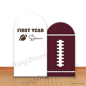 May include: Two arched backdrop banners. One white banner with the text "FIRST YEAR Down" and a football graphic. The other banner is maroon with a white football lace design. The banners are for a football-themed event.