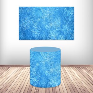May include: A blue cylinder-shaped display stand with a frosted snowflake pattern. The stand is on a wooden floor.