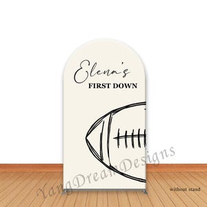 May include: An off-white arched sign with the text "Elena's FIRST DOWN" in black script. A black line drawing of a football is below the text. The sign is on a metal stand.