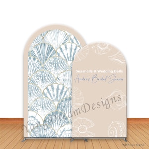 May include: Two arched backdrop banners. One features a blue and white seashell pattern with gold accents. The other is beige with seashell illustrations and text that reads "Seashells & Wedding Bells, Andre's Bridal Shower."