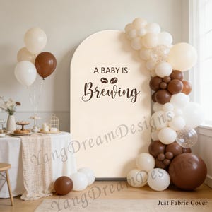 May include: A baby shower backdrop with the text "A Baby Is Brewing" in brown script. The backdrop is decorated with a balloon arch in white, brown, and clear balloons. Balloons in similar colours are also scattered around the room.
