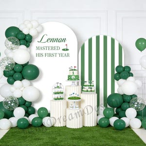 Könnte beinhalten: Eine Party zum ersten Geburtstag mit Golf-Motiv, mit grünen und weißen Luftballons, Kuchen und Cupcakes. Der Hintergrund zeigt einen weißen Bogen mit dem Text "Lennon Mastered His First Year". Die Szene umfasst einen grünen Rasenboden.