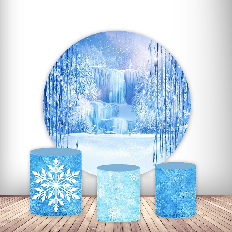 May include: Three blue and white cylindrical cardboard cake stands with a snowflake design. The background is a round backdrop with a winter wonderland scene featuring a frozen waterfall and trees.