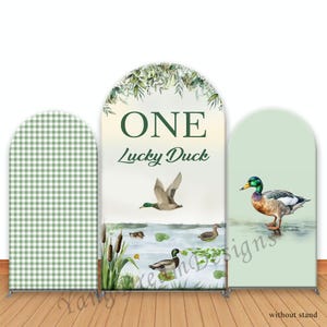 Peut inclure: Trois panneaux de fond arqués. Le panneau central présente une scène à l'aquarelle avec des canards, des quenouilles et le texte "ONE Lucky Duck". Le panneau de gauche a un motif vichy vert et blanc. Le panneau de droite montre un canard dans l'eau.
