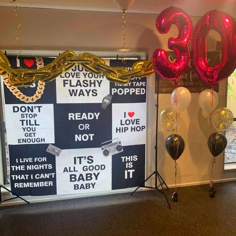 May include: A black and white backdrop with a 90s theme for a 30th birthday party. The backdrop features phrases like "Love Your Flashy Ways", "Ready or Not", "I Love Hip Hop", and "It's All Good Baby Baby". The backdrop is decorated with a gold chain garland and a large pink number 30 balloon.