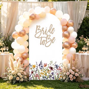 May include: A white arched backdrop featuring the text Bride to Be in a script font. The base is decorated with a colorful floral print of wildflowers. The arch is framed by a balloon garland in shades of white, cream, and metallic copper.