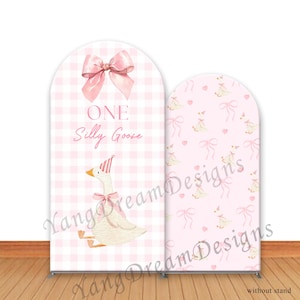 Peut inclure: Deux bannières de fond arquées. L'une présente un motif vichy rose avec un nœud rose et le texte "ONE Silly Goose" et une oie portant un chapeau de fête. L'autre bannière a un motif rose avec des oies et des nœuds.