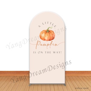 May include: A light peach-colored banner with an arched top. The banner features a watercolor illustration of an orange pumpkin and the text "A LITTLE Pumpkin IS ON THE WAY!" in a script font. The words "Yang Dream Designs" are also visible.