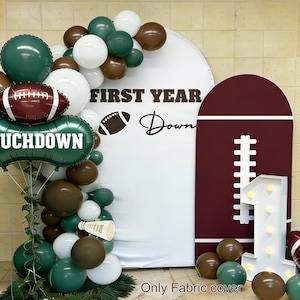 Könnte beinhalten: Dekoration für eine Fußball-Motto-Geburtstagsfeier. Enthält einen weißen Bogen mit dem Text "FIRST YEAR Down", einen weinroten Football-förmigen Hintergrund und eine beleuchtete Zahl eins. Ballons in Grün, Weiß und Braun vervollständigen den festlichen Look.