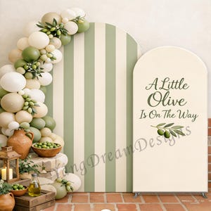Peut inclure: Un décor de fête avec un fond à rayures crème et vert olive, et une pancarte avec l'inscription "A Little Olive Is On The Way". La scène est ornée de ballons, d'olives et de bouteilles d'huile d'olive.