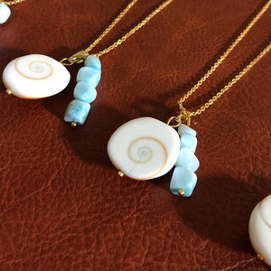 May include: Five gold necklaces with a white shell pendant and blue beads. The necklaces are displayed on a brown leather surface.