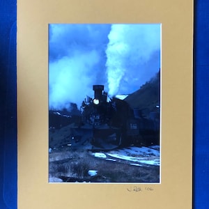 May include: A black steam locomotive with a plume of white smoke billowing from its stack, travelling through a snowy mountain landscape. The locomotive is labelled "1455" and "Rio Grande" on its side. The image is signed "J. Roth '06" in the bottom right corner.