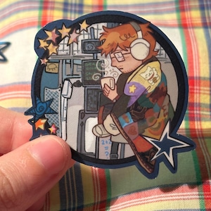 May include: A die-cut sticker featuring an illustration of a person wearing headphones, glasses, and a colorful patchwork jacket, holding a mug. The background includes screens and stars. The sticker is held by a hand.