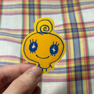 K&ouml;nnte beinhalten: Ein gelber Aufkleber mit einer Cartoonfigur mit blauen Augen und einem l&auml;chelnden Mund. Die Figur hat eine Spirale auf dem Kopf und ist blau umrandet. Der Aufkleber wird von einer Hand gehalten.
