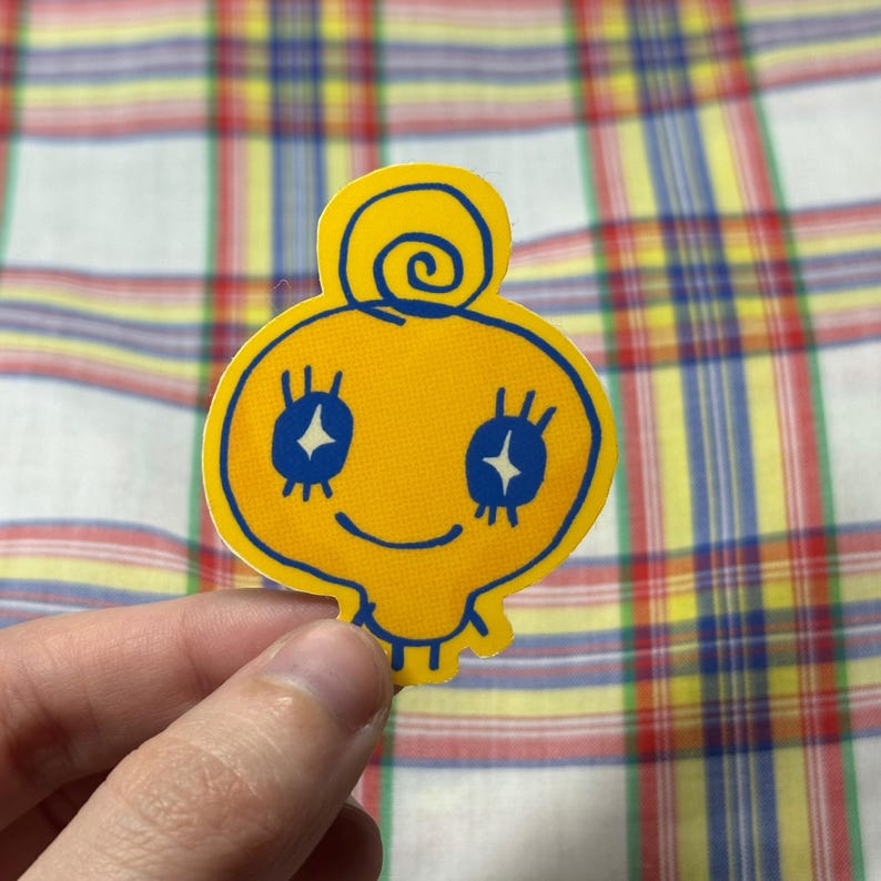 K&ouml;nnte beinhalten: Ein gelber Aufkleber mit einer Cartoonfigur mit blauen Augen und einem l&auml;chelnden Mund. Die Figur hat eine Spirale auf dem Kopf und ist blau umrandet. Der Aufkleber wird von einer Hand gehalten.