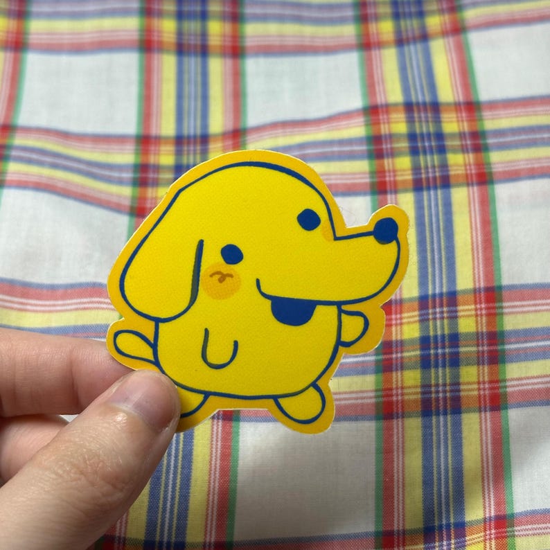 K&ouml;nnte beinhalten: Ein gelber Aufkleber mit einem Cartoon-Hund mit blauem Umriss. Der Hund hat einen l&auml;chelnden Ausdruck, mit blauem Mund und Augen und einer pfirsichfarbenen Wange. Der Aufkleber wird vor einem karierten Stoffhintergrund gehalten.