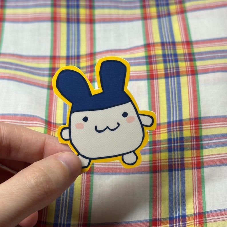 K&ouml;nnte beinhalten: Ein gestanzter Aufkleber mit einem Cartoon-Hasencharakter mit wei&szlig;em K&ouml;rper, blauem Kopf und gelbem Umriss. Der Hase hat ein l&auml;chelndes Gesicht mit kleinen schwarzen Punkten f&uuml;r die Augen und einem gebogenen Mund.