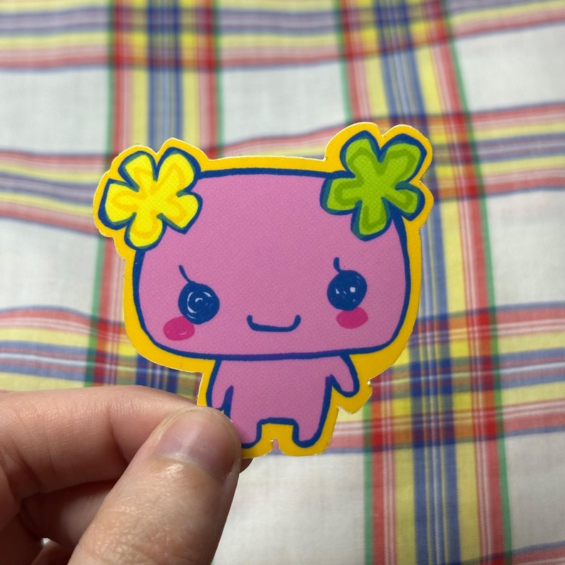K&ouml;nnte beinhalten: Ein pinkfarbener Aufkleber mit einer Cartoonfigur mit gelbem Umriss. Die Figur hat blaue Augen, rosa Wangen und gelbe und gr&uuml;ne bl&uuml;tenf&ouml;rmige Haaraccessoires. Der Aufkleber wird vor einem karierten Hintergrund gehalten.