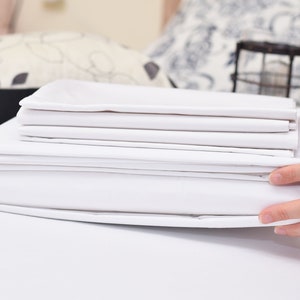 May include: A stack of folded white bed sheets, including a fitted sheet, flat sheet, and pillowcases. The sheets are neatly arranged and held by a person's hands. The bedding is crisp and clean, suggesting a fresh and inviting bedroom.