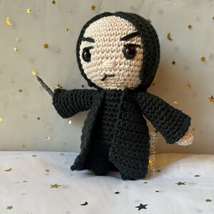 Può includere: Bambola all'uncinetto di un personaggio con abito nero, cappuccio nero e viso pallido. La bambola ha grandi occhi neri e tiene in mano una bacchetta. Lo sfondo è un tessuto chiaro con dettagli di stelle e lune dorate.